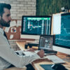 Businessman in a white shirt sits at a desk with multiple monitors displaying stock charts and trading data.