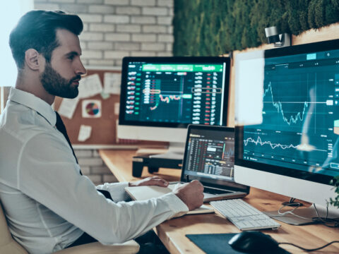 Businessman in a white shirt sits at a desk with multiple monitors displaying stock charts and trading data.