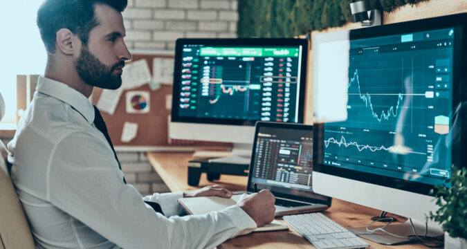 Businessman in a white shirt sits at a desk with multiple monitors displaying stock charts and trading data.