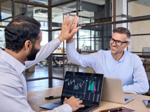 Two colleagues in a bright office high-five over a laptop displaying stock charts and data.