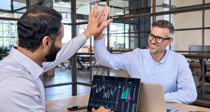 Two colleagues in a bright office high-five over a laptop displaying stock charts and data.